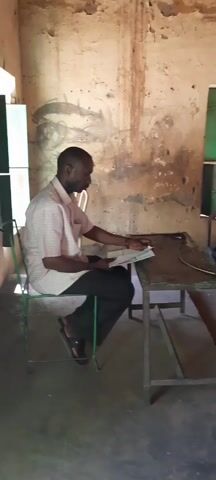 The video shows a classroom of students, all dressed in light green shirts and dark pants. They are seated at desks in rows. An adult male, dressed in a light pink shirt and dark pants, sits at a desk in the front and appears to be reading from a document. The walls of the classroom are off-white and appear old or unfinished, with visible wear and some markings. There is at least one green-framed window in the room.
