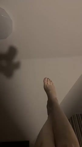 The video shows a pair of bare human feet and legs from a low-angle perspective, pointing upwards towards a ceiling. The feet are positioned with the toes facing the viewer, and the legs are bent at the knees, creating an inverted V-shape. The scene is dimly lit, with a shadow of what appears to be a light fixture visible on the ceiling. The background includes a hint of a striped pattern, suggesting furniture or a wall covering. The feet and legs are the primary focus, and they appear to be still for the most part, with subtle movements.