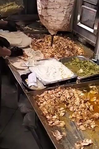 The video shows a close-up of a food preparation area, possibly a restaurant kitchen. A large vertical rotisserie of meat is the central feature. In front of it are trays containing sliced and cooked meat, a container of white sauce (likely tzatziki or a similar yogurt-based sauce), and a tray of sliced green vegetables. A person wearing black gloves is spreading white sauce on a flatbread, presumably to make a wrap or sandwich. The surface is stainless steel and appears to be in a well-used state.