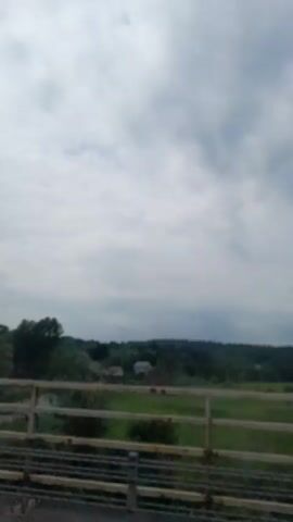 The video captures a scenic view from a bridge overlooking a rural landscape under a cloudy sky. In the foreground, a wooden railing of the bridge is visible, suggesting a vantage point for observing the scene below.

The main subject of the scene is a winding road that cuts through lush green fields and dense trees. A white car is seen traveling along the road, indicating a sense of movement and activity within the otherwise tranquil setting. In the distance, a small white house with a red roof is nestled amongst the trees, adding a touch of habitation to the natural environment. The trees are varied in type and size, with some appearing more mature and others younger, contributing to the textured appearance of the landscape.

The weather appears to be overcast, with a dramatic sky filled with dark and light clouds. This suggests a possibly humid or impending rain. The time of day is indeterminate due to the cloud cover, but the overall lighting suggests it could be daytime. The location is identified as Balice, Poland, which places the scene in a European rural setting. The atmosphere evoked is one of peace and natural beauty, with the presence of the road and car hinting at a subtle human presence within the natural environment. There is no visible text in the video.