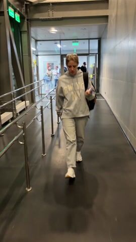 A young woman walks along an airport jet bridge toward the camera. She wears a gray hooded sweatshirt with "NEW YORK" printed on it, gray sweatpants, and white sneakers. She also wears a tiara and carries a backpack. The jet bridge is metal and gray, with a dark gray floor and silver handrails. Other passengers can be seen in the background.