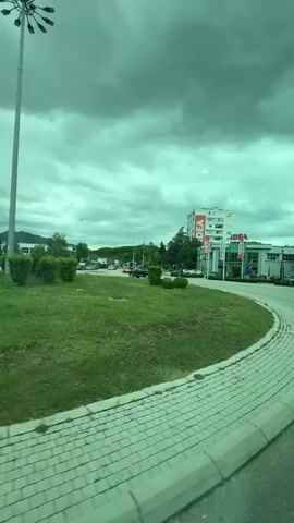 A video taken from inside a moving vehicle shows an intersection in what appears to be a town. The sky is overcast, with a green tint to the entire scene, possibly due to a tinted window or camera filter. In the background, there is a multi-story building with "IDEA" signage prominently displayed. The intersection has a roundabout with a grassy center and a pedestrian crosswalk. A person wearing an orange jumpsuit and carrying a suitcase is seen crossing the road. Cars are visible on the streets. A billboard is visible advertising an event featuring “MUCCI”. The street is lined with trees and streetlights.