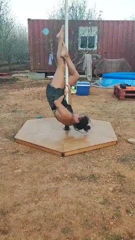 A woman performs a pole dancing routine outdoors on a hexagonal platform. She is wearing a black bikini top and denim shorts. The background includes a red shipping container, trees, and a blue inflatable pool. The ground is dirt.