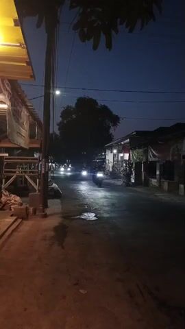 The video shows a dimly lit street at dusk or night. A motorcycle with its headlights on is the most prominent moving object, heading away from the camera and illuminating the wet asphalt road. To the left, a building with a yellow awning and some stacked materials is visible. On the right, several buildings with lights are lined up along the street. The sky is a deep blue, transitioning into darkness. A tree is silhouetted against the sky in the distance. The overall atmosphere is quiet and somewhat isolated.