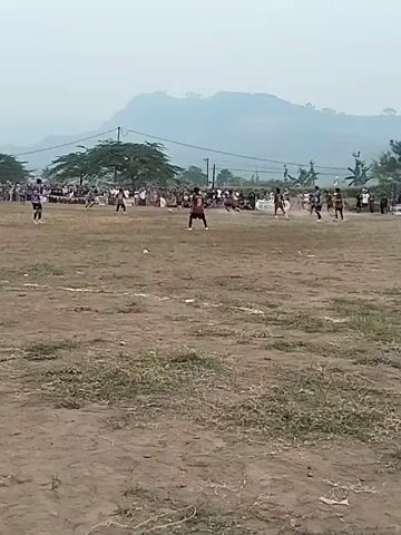 A group of people are playing soccer on a dirt field. There are spectators in the background. The sky is hazy and there are mountains in the distance. One player in a red jersey is dribbling the ball.