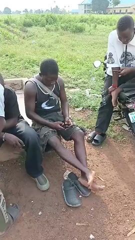 A group of four young Black men are gathered outdoors. Two men are sitting on a low concrete structure, facing each other and looking at their phones. One man, wearing a black tank top with a white logo and dark shorts, is in the foreground, with his legs crossed. Another man, wearing a black t-shirt and dark pants, is sitting next to him. A third man, wearing a light grey t-shirt and green cargo pants, stands behind them, also looking at his phone. A fourth man, wearing a black and white patterned shirt and dark pants, stands on the right side of the frame, mostly out of view. The ground is dirt and gravel, with some sparse vegetation in the background. A white motorcycle is partially visible on the right side of the image. The sky is overcast.