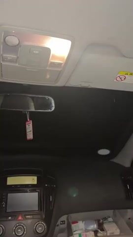 The video shows the interior of a car, specifically the front portion of the passenger cabin. The camera is positioned from the driver's seat, looking up at the ceiling and dashboard. The cabin light is on, illuminating the overhead console and surrounding area. A rear-view mirror is visible, hanging down. The dashboard includes a radio/navigation system, climate control knobs, and some miscellaneous items stored in a compartment below. At second zero, a white object is thrown toward the camera. At second one, the sound of a buzzer is heard.