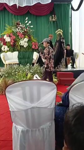 The video shows a wedding ceremony. A man is filming the event with a camera. A couple, dressed in traditional wedding attire, is seated on a stage decorated with flowers. The groom is wearing a brown patterned sarong and a black top with a headpiece. The bride is wearing a black dress with intricate embroidery and a veil, also with a headpiece. Several other individuals are present on the stage, some of whom appear to be offering blessings or assistance to the couple. The stage is set against a backdrop of green curtains, with additional floral arrangements and white chairs. In the foreground, several chairs are covered with white fabric. The overall atmosphere appears to be formal and celebratory.