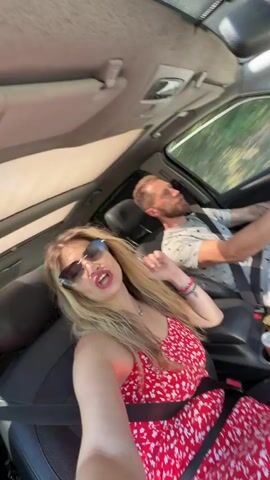 A cheerful blonde woman wearing sunglasses and a red floral dress is seen in the front passenger seat of a car, buckled up. She is actively singing along to music, smiling, and moving her hands expressively. A man with a beard is driving the car, also wearing a seatbelt and a light-colored T-shirt with a pattern. He appears focused on the road but has a relaxed expression, occasionally looking forward and to the side.

The car is moving, indicated by the blurred scenery outside the windows. The sun visor is up on the passenger side, and there are a couple of cans in the cup holders in the center console. The interior of the car is visible, showing the dashboard and parts of the ceiling. The overall atmosphere is joyful and relaxed. The scene takes place during the daytime, with bright natural light coming through the windows. The activity suggests a road trip or casual drive. The specified location for this scene is Medinski Krš, Montenegro.