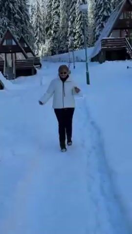 A woman wearing a white jacket, dark pants, and brown shoes walks down a snowy path between A-frame chalets. She is smiling and has her arms slightly outstretched. In the background, snow-covered fir trees reach into a clear blue sky. A baby stroller is visible near one of the chalets. The scene appears to be a winter resort or village.