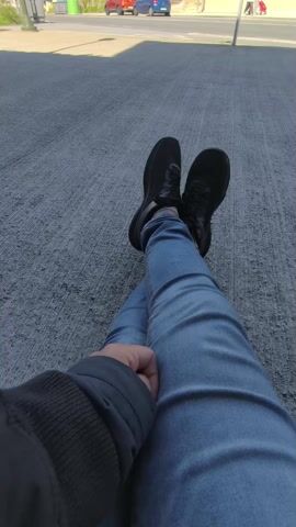 The video presents a first-person perspective from someone resting outdoors in Cangas de Morrazo, Spain. The viewer's legs are extended and crossed at the ankles, clad in light blue jeans and black athletic shoes. A portion of a dark-colored jacket or sleeve is visible on the left side of the frame, belonging to the person filming. The immediate ground beneath is a textured grey concrete surface.

In the background, a bright, sunny street scene unfolds under a clear blue sky, indicating daytime. Buildings with varying architectural styles and colors, including a prominent white and light green structure and another with an orange-brown roof, line the street. Several cars are visible on the road, including a red car and a blue car. A traffic light and streetlights are also present, confirming an urban or suburban setting. In the distance on the sidewalk, two small figures can be seen walking, one appearing to push a red cart. No discernible text is visible anywhere in the video. The overall impression is one of a calm, everyday street scene during good weather.