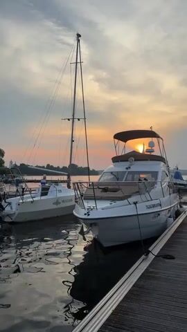 The video shows a harbor with several boats docked at sunset. The sky is cloudy with a warm orange glow from the setting sun. The foreground is dominated by the reflections of the boats and the sky on the water. On the right, a white motor yacht is docked, its stern facing the camera. To its left, a white sailboat is also docked, with its mast extending high into the sky. Further in the background, more boats are visible, as well as trees and a building on the shore. The overall impression is one of peacefulness and natural beauty.