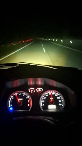 The video displays a first-person perspective from the driver's seat of a car traveling on a highway at night. The dashboard is visible in the foreground, featuring a speedometer on the right and a tachometer on the left, both illuminated with red and white lighting. The speedometer needle indicates acceleration, moving past 90 km/h and approaching 100 km/h. Bright headlights illuminate the multi-lane road ahead, which is marked with white lines. Red reflective markers are visible along the left side of the highway, while white or yellow markers are visible on the right.