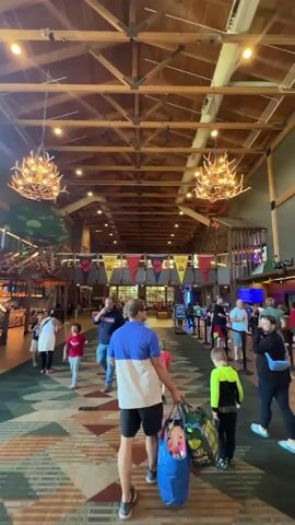 The video shows a busy indoor lobby with a high, wooden beam ceiling and decorative antler chandeliers. A patterned carpet covers the floor. In the foreground, a man in a blue and white shirt carries several large bags as he walks away from the camera, followed by two children. Other people are walking in various directions throughout the lobby. In the background, there appears to be a reception or check-in area with a queue of people. Colorful banners with paw print logos are hanging from the ceiling. The overall impression is a lively, family-friendly environment.