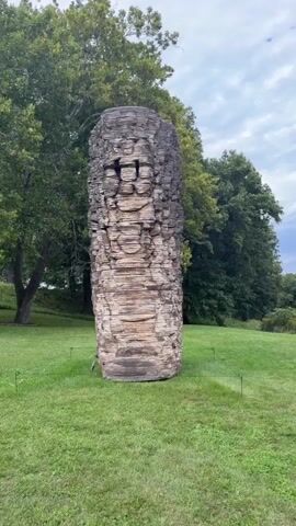 The video shows a tall, cylindrical stone sculpture situated in a grassy field with trees in the background under a cloudy sky. The sculpture is made of stacked, irregularly shaped stones, giving it a rough, textured appearance. A thin metal pole is visible attached to the left side of the sculpture near its base. Small, dark stakes are placed in the grass around the sculpture, marking its perimeter. The camera pans slightly upward, revealing more of the sculpture's height and the surrounding trees.