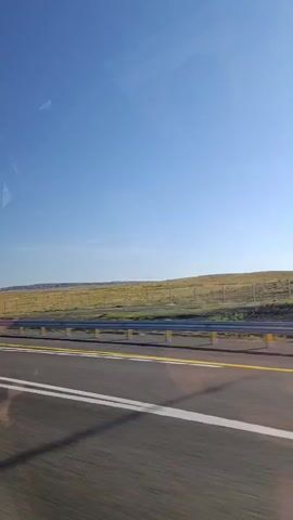 The video shows a view from a vehicle traveling on a highway. The sky is clear blue. On the right side of the road, there is a field of dry grass and a guardrail. A street lamp is visible on the left side of the road. A tow truck carrying another vehicle passes by. The video then returns to the view of the highway and the field.