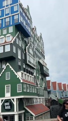 The video shows a unique hotel building with an unusual architectural design that resembles a stack of traditional Dutch houses. The building is predominantly green, with sections in blue and white, and features a stepped design with overhanging sections. The houses have pointed gables, numerous windows, and red tiled roofs.

At the base of the building, a sign displays "Inntel Hotels" in white text within a black circle. Below this, another sign reads "Food & Drinks". A small portion of a person's arm holding a phone is visible in the bottom left corner, suggesting someone is filming or taking pictures. A woman with dark hair and wearing a dark t-shirt is partially visible on the right side of the frame.

In the background, several other buildings with similar Dutch architectural styles can be seen, some with red roofs and white trim. A bridge or walkway is also visible connecting some of these structures. The sky is overcast, suggesting a cloudy day. The overall scene depicts a striking and whimsical hotel that is a prominent landmark, likely in Zaandam, Netherlands, given the architectural style. The atmosphere appears to be one of curiosity and appreciation for the distinctive design.
