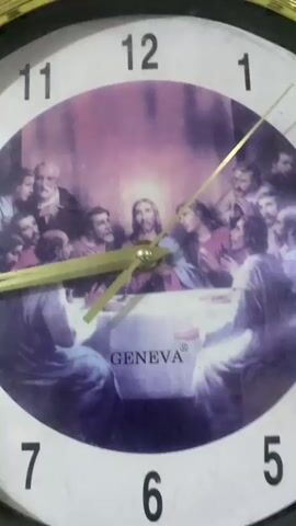 This media file is a close-up, slightly angled shot of a round analog clock. The clock face features a circular depiction of Leonardo da Vinci's famous fresco, "The Last Supper," which shows Jesus Christ and his twelve apostles gathered around a table. The painting is rendered in a purple-hued, somewhat muted style, with a glowing aura around Jesus's head.

The clock has traditional hour markings from 1 to 12, with the numbers themselves in black. Two golden clock hands are visible. The hour hand is shorter and thicker, pointing slightly past the 4. The minute hand is longer and thinner, and it points directly at the 12. This indicates that the time displayed on the clock is approximately 4:00.

Below the painting, the word "GENEVA" is visible in a white, stylized font, with a small registered trademark symbol (®) to its right. The clock itself appears to be framed by a dark, possibly black or dark brown, material. The background is a blurred mixture of light and dark tones. The overall impression is of a decorative clock, possibly intended for a home or religious setting, with the iconic "Last Supper" scene serving as its visual centerpiece. The image quality suggests it was taken with a handheld device, possibly a mobile phone, in natural light conditions.