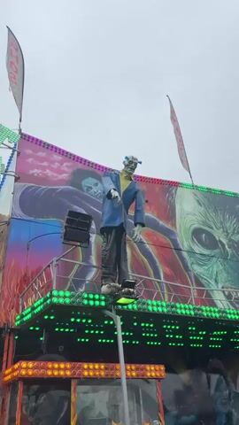 A slow pan of a carnival ride with a horror theme. The ride is decorated with images of monsters and skulls. A large, detached zombie figure stands on a platform at the top of the ride. A large, green-skinned alien-like creature is painted on the side of the ride. The ride is lit with green and orange lights. People can be seen in the background. The sky is overcast.