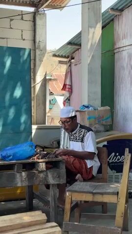 A man wearing a white kufi and glasses is sitting at a wooden table. He is wearing a white shirt with a black and white patterned yoke and orange sarong. He is holding a small, dark brown object in his hands and appears to be working on it with a tool. Several similar objects are lined up on the table in front of him, along with a blue plastic bag. The table is set up outdoors, with a building in the background that has corrugated metal roofing and colorful siding. Sunlight is casting shadows.