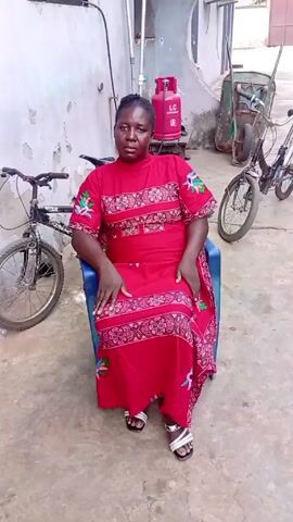A woman is sitting in a blue plastic chair in an outdoor, enclosed space. She is wearing a red dress with pink and green patterns, and white sandals. Her hair is pulled back. Behind her, there is a white wall with pipes, a red gas cylinder, a green wheelbarrow, and two bicycles. The ground is concrete. The woman is looking directly at the camera. Someone says, "Good morning, sir."