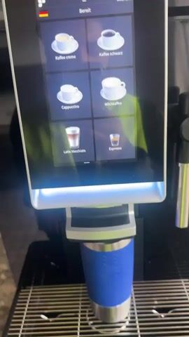 A person's hand with dark skin and short fingernails is visible in the foreground. The hand is positioned over a blue insulated travel mug that is sitting on the drip tray of a coffee machine. The coffee machine has a large touchscreen display. The display shows a menu of coffee options, including "Kaffee Creme", "Kaffee schwarz", "Cappuccino", "Milchkaffee", "Latte Macchiato", and "Espresso". The person's index finger is touching the "Espresso" option on the screen. The coffee machine then displays a message that says "Getränk wird zubereitet..." and shows a progress bar at 0%. The background is blurry but appears to be an indoor environment with a bright yellow or green object behind the coffee machine.