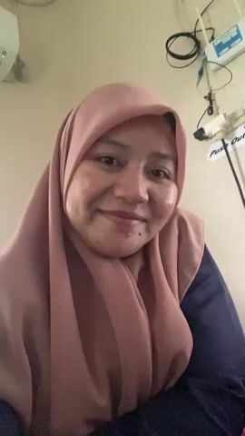 The video shows a close-up of a young woman wearing a pink hijab and a dark blue top. She is indoors, in what appears to be a room with medical equipment on the wall behind her. The woman smiles and turns her head slightly. The lighting is warm and somewhat dim.
