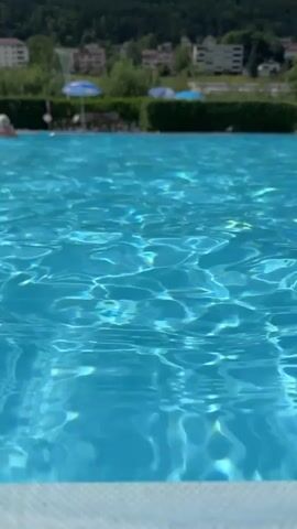 This is a close-up, low-angle shot of the surface of a bright blue swimming pool. The water is slightly rippled, with sunlight reflecting off the surface, creating shimmering patterns. In the blurred background, there are hints of a green hedge, some buildings on a hillside, and a few people in and around the pool. The foreground shows the white edge of the pool. The overall impression is one of a sunny day at a public swimming facility.