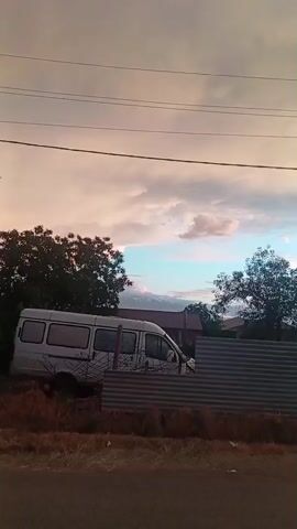 This is a video clip showing a street scene at dusk in Vedi, Armenia. The sun is setting, casting a warm glow on the sky, with hues of pink, orange, and blue visible in the clouds. A white van is parked on the left side of the frame, partially obscured by a corrugated metal fence. Behind the van, trees and houses can be seen. A power pole with numerous wires stretches across the sky. The overall atmosphere is peaceful and serene, indicative of the end of the day. No people are visible in the frame.
