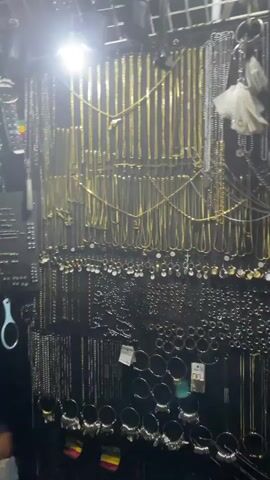 The video starts with a close shot of a dense display of jewelry, predominantly chains and earrings, hanging against a dark backdrop. The jewelry is predominantly gold and silver colored, with some black pieces. A bright light illuminates the display. The camera then pans upward, showing more of the display rack and overhead lighting. Finally, the camera angle shifts abruptly, showing foliage in a very dark setting, with hints of colored lights in the background. There are no discernible people in the frame.
