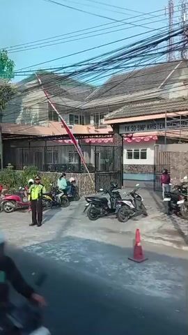 The video shows a street scene with several motorcycles. In the foreground, a green motorcycle with two riders passes by. Behind them, a white motorcycle with one rider is visible. To the left, a red motorcycle with two riders is parked. Further back, a white motorcycle is parked, and a man in a red shirt stands beside it. Another man, wearing a yellow jacket, is on a motorcycle in front of a building with a fence. The building has a green sign that reads "SEKOLAH KRISTEN PAMERDT TK SD SMP" and a flag with red and white stripes is flying on a pole. Trees and power lines are visible overhead. The overall environment appears to be an urban or suburban area on a sunny day.