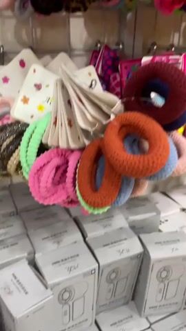 The video pans across a display of hair accessories, primarily scrunchies and hair ties, in various colors and patterns. The items are neatly arranged on shelves and hanging displays. Boxes of what appear to be retail merchandise are stacked on the floor in the foreground. The lighting suggests an indoor retail environment, possibly a shop or market stall. There are no people visible in the frame.