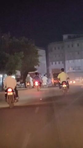 Two people on a motorcycle drive down a street at night. The person in the back is wearing a blue shirt and the person in the front is wearing a white shirt. A second motorcycle with two people on it is in front of them. The person in the back is wearing a white shirt. The person in the front is wearing a light blue shirt. Buildings are visible on the right side of the street.