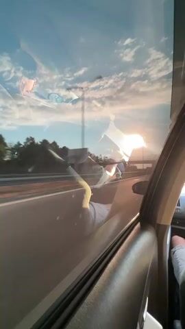 The video shows a view from inside a car, looking out the passenger window. The sun is setting or rising, casting a warm glow and creating lens flares on the window. There are reflections on the window, including what appears to be a person sitting in the passenger seat, with their legs extended. Outside the car, there is a highway with trees and some structures visible in the distance. The car is in motion, as indicated by the blurred scenery.