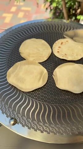 The media file captures a close-up, overhead view of four flatbreads being cooked on a griddle. The flatbreads are in various stages of cooking, with some appearing pale and doughy, while others have begun to brown and develop small blisters. Tongs are visible, suggesting someone is actively managing the cooking process. The griddle itself has a distinctive patterned surface with a honeycomb design and wavy lines. In the background, a blurred image of a paved area with yellow and orange squares suggests an outdoor setting, possibly a patio or courtyard. Sunlight illuminates the scene, indicating it's daytime. The overall impression is one of a domestic cooking activity, likely in a warm, possibly sunny climate given the outdoor setting and the type of food being prepared. No text is visible in the media file. The location context provided is Even Yehuda, Israel.