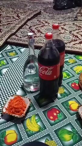 The video shows a low table setting on the floor, likely for a meal. A patterned rug is visible beneath the table setting. On the table, which has a checkered and fruit-themed tablecloth, there are two large bottles of Coca-Cola, a glass bottle filled with clear liquid (possibly water or a soft drink), and a small square dish filled with shredded orange carrots. The table is positioned in what appears to be a room setting. A dark bag is visible in the background, indicating a room environment. There are no people fully visible in the frame, but a small portion of black clothing is present at the bottom, suggesting the presence of a person behind the camera.