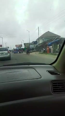 The video, filmed from inside a car, shows a street scene with moderate traffic. A silver car is in front of the camera, followed by several colorful auto-rickshaws. On the right side of the road, there are buildings that appear to be shops, and a few people are visible walking and standing near them. The sky is overcast. The camera is stationary throughout the short clip.