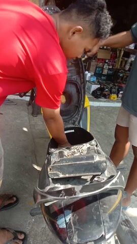 The video shows two men working on a black motorcycle. The man in the foreground, wearing a red t-shirt, is leaning over the motorcycle, appearing to be working on the engine or a part of the motorcycle's body. He has his left hand inside an open compartment of the motorcycle. To his right, another man, wearing a dark grey t-shirt and light-colored shorts, is also leaning over the motorcycle, his hands on it. In the background, other people and a yellow motorcycle are visible. The environment appears to be an outdoor, possibly a street or a shop area, with a concrete ground. There are some shelves or displays with various items in the background. The lighting suggests it is daytime.