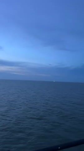 This is a video clip taken from a boat, looking out over the ocean. The sky is a deep blue, indicating it is either dawn or dusk. There are scattered clouds, and the water is calm with gentle ripples. In the distance, a faint line of land is visible, possibly a coastline or islands.

There are no people visible in the foreground. In the middle ground, a faint light can be seen on the horizon, likely from a distant boat or structure. The foreground shows a dark railing, part of the boat from which the video is being filmed. A small flag, with red, white, and blue stripes, is attached to a pole on the right side of the frame. The flag appears to be the Thai flag.

The overall mood is peaceful and serene, suggesting a calm moment at sea. The location cues, particularly the Thai flag, point to a setting in Thailand, specifically near Thong Nian as provided. The time of day is either early morning or late evening, as suggested by the deep blue sky. There is no visible text in the video.