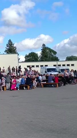 A crowd of people, mostly adults and some children, are gathered outdoors in front of a low, white building with a light blue trim. They are seated on benches or standing. The ground is paved with grey asphalt. Trees and a white tent are visible behind the crowd. The sky is blue with scattered white clouds. The camera is panning from left to right.