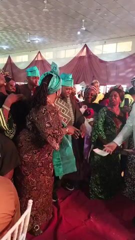 The video captures a vibrant indoor celebration, likely a wedding reception, set in Fagge D2, Nigeria. Numerous adults, predominantly dressed in traditional Nigerian attire, are gathered on a red carpet. In the foreground, a woman wearing a brown patterned dress and a teal headwrap is seen moving gracefully, facing a group of smiling men and women. Many attendees are adorned with matching teal headwraps or caps, signifying a unified theme. A man in a light-colored, long tunic and a patterned cap stands on the right, holding what appears to be money, with banknotes scattered across the red carpet, indicating the cultural practice of money spraying during celebrations. The background features decorative burgundy drapes and ceiling fans, suggesting an event hall or banquet space. The atmosphere is joyful and festive, with visible smiles and expressions of happiness among the participants.