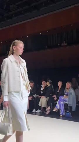 The video shows a fashion show. A female model with long blonde hair walks down a runway wearing a white tweed jacket and a white skirt. She is carrying a white handbag with a pearl strap. Several people are seated in the audience, watching the show. The environment appears to be a theater or auditorium.