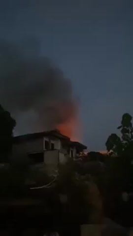 The video shows a two-story building engulfed in fire with thick, black smoke billowing from behind it. The sky is a dusky blue, suggesting it is either dawn or dusk. The fire is visible rising above the roofline, indicating a significant blaze. The building appears to be situated on a slight incline or hill, with some vegetation visible in the foreground. The overall scene is one of destruction and emergency.