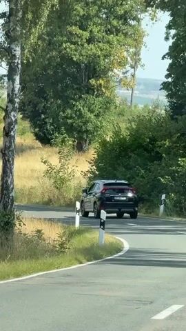 A black Mitsubishi Eclipse Cross drives on a winding asphalt road in a rural setting with dry grass fields and trees. A motorcycle rider in a helmet and riding gear, on a black and silver motorcycle with saddlebags, approaches and passes the SUV. The sun is shining.