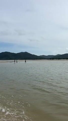 A vertical shot shows a body of water with several people in it. In the foreground, the water is shallow, and the sandy bottom is visible. Small waves are breaking on the shore. In the middle ground, several people are standing in the water, some appearing to be wading or paddleboarding. In the background, a low mountain range is visible under a cloudy sky. The overall impression is a calm, natural scene.
