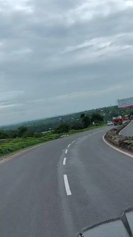 The video shows a view from inside a vehicle traveling on a paved road. The road is gray asphalt with white dashed lines marking the lanes. The vehicle appears to be in the right lane of a highway, and there are other vehicles on the road, including trucks and cars. The landscape outside the vehicle is green with trees and vegetation. The sky is overcast with gray clouds. There are billboards visible in the distance on the right side of the road. The perspective is from a low angle, looking forward and slightly to the right.