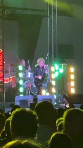 A man with blond hair and a black jacket is singing on stage. There are lights behind him and a crowd in front of him. A phone is in the foreground, recording the concert.