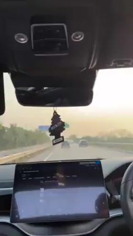 The video shows the interior of a car driving on a highway. The car's dashboard features a large touchscreen display, and a "Little Trees" air freshener hangs from the rearview mirror. The road ahead has multiple lanes with a few other cars visible in the distance. The sky is overcast, creating a soft light, with trees lining the side of the road.