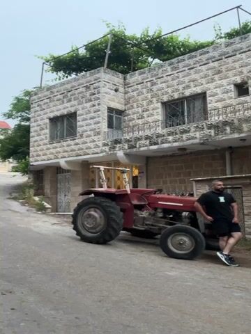 A red tractor is parked on the side of a paved road in front of a building with stone siding. The building has at least two windows and a vine growing on its roof. Trees and other buildings are visible in the background. The sky is overcast. The video is shot from a low angle, looking up at the tractor and building.