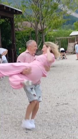 A man in a pink plaid shirt and blue shorts is carrying a woman in a pink jumpsuit. They are outdoors in a gravel area with trees and mountains in the background. Other people are visible in the background. The man is holding the woman in his arms, and she appears to be laughing. They are in motion, possibly dancing or spinning.